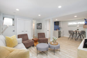 Renovated Edmonton basement living area with white walls, light floors, 2 brown lounge chairs, grey ottomans and closet view.