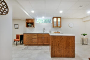 Renovated basement bar with a white tile wall, sink, brown cabinets and two cupboards with a small window in between.