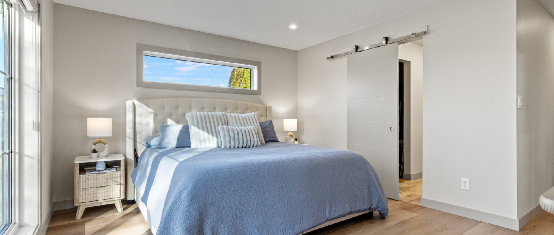 Bright modern bedroom with a large bed dressed in blue and white linens. The room has light wood flooring, soft lighting, and a small window.