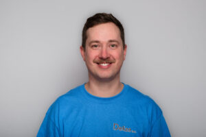 A headshot of a man with short dark blonde hair and a moustache up against a grey background wearing a blue t-shirt