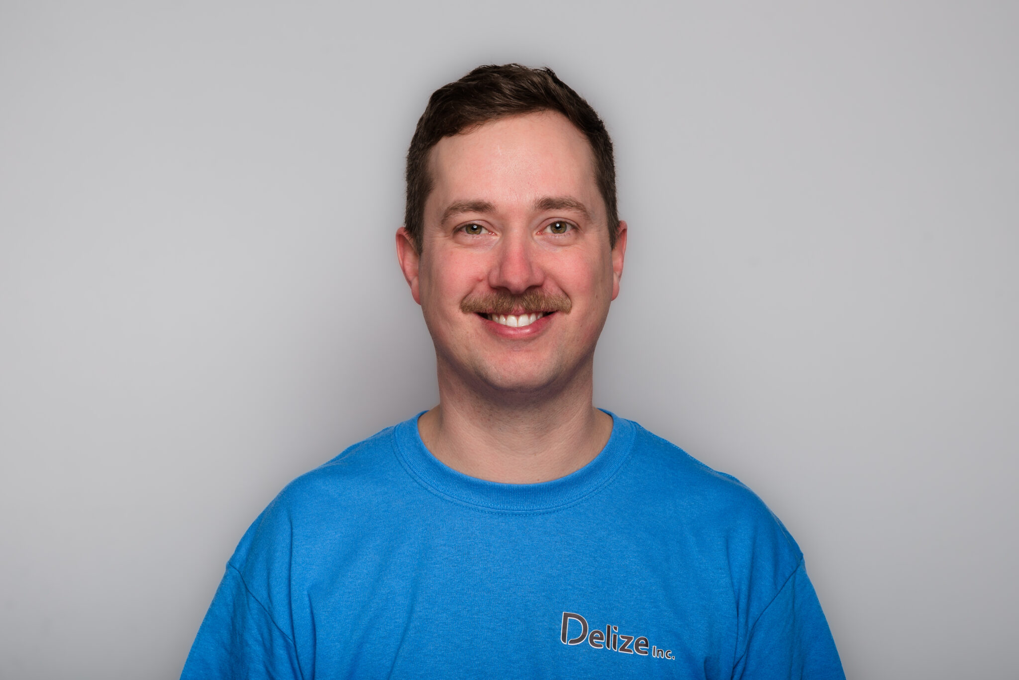 A headshot of a man with short dark blonde hair and a moustache up against a grey background wearing a blue t-shirt