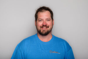 A headshot of a man with short dark hair and beard. He's wearing a blue t-shirt and is up against a grey background.