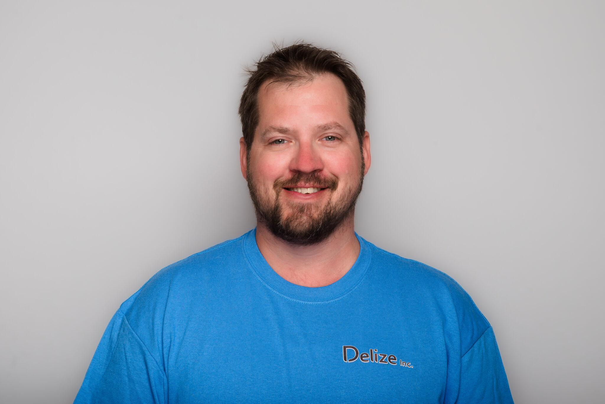 A headshot of a man with short dark hair and beard. He's wearing a blue t-shirt and is up against a grey background.