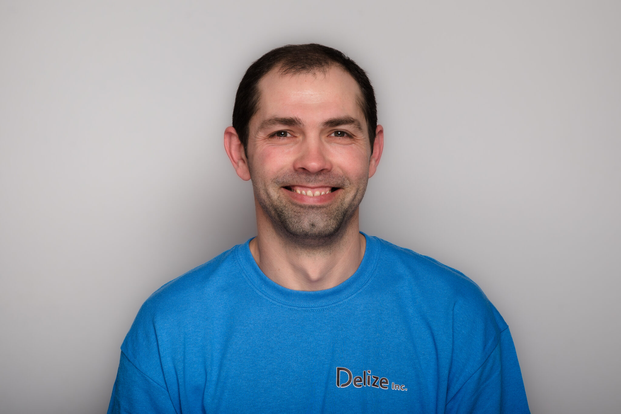 A headshot of a smiling man with short dark hair. He's wearing a blue t-shirt and is photographed against a grey wall.