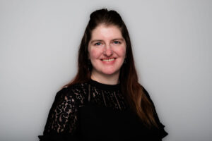 A headshot of a woman with long dark hair, wearing a black blouse with lace accents. She is photographed against a grey background.