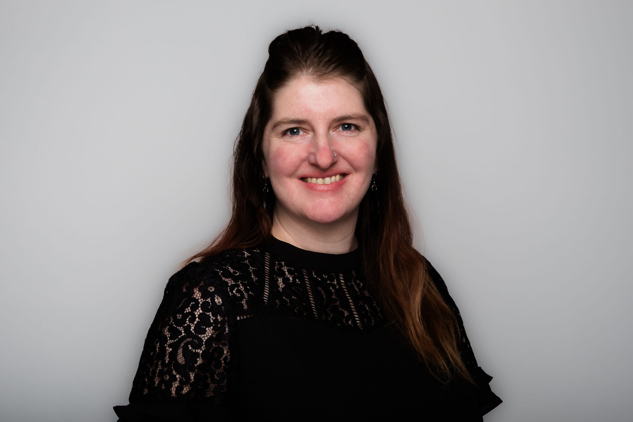 A headshot of a woman with long dark hair, wearing a black blouse with lace accents. She is photographed against a grey background.