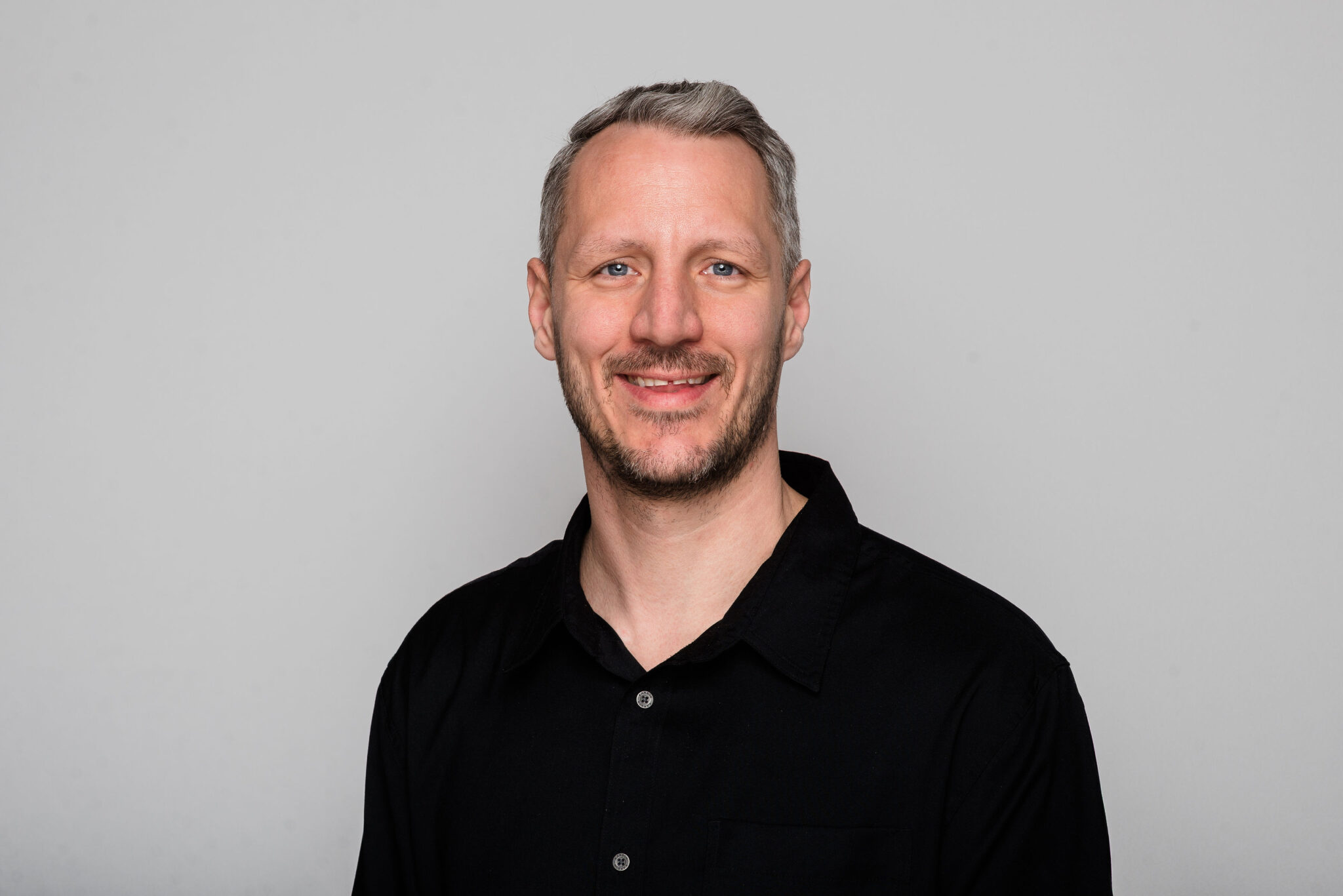 A headshot of a man with short grey and dark blonde hair wearing a black polo style shirt.