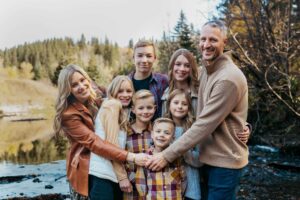 A portrait of a family of 8. A mom and dad, 3 girls and 3 boys. The photo was taken in the woods. There are trees in the background.