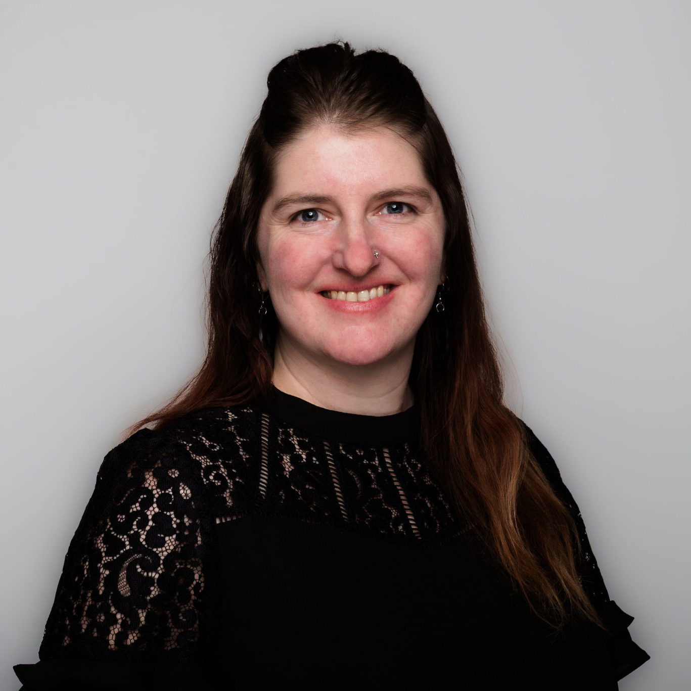 A headshot of a woman with long dark hair, wearing a black blouse with lace accents. She is photographed against a grey background.